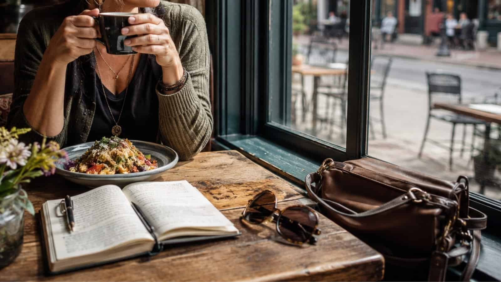 A woman taking a photo of her meal at a cozy café window, with a notebook, sunglasses, and a bag on the table, capturing the essence of solo travel and enjoying local experiences.