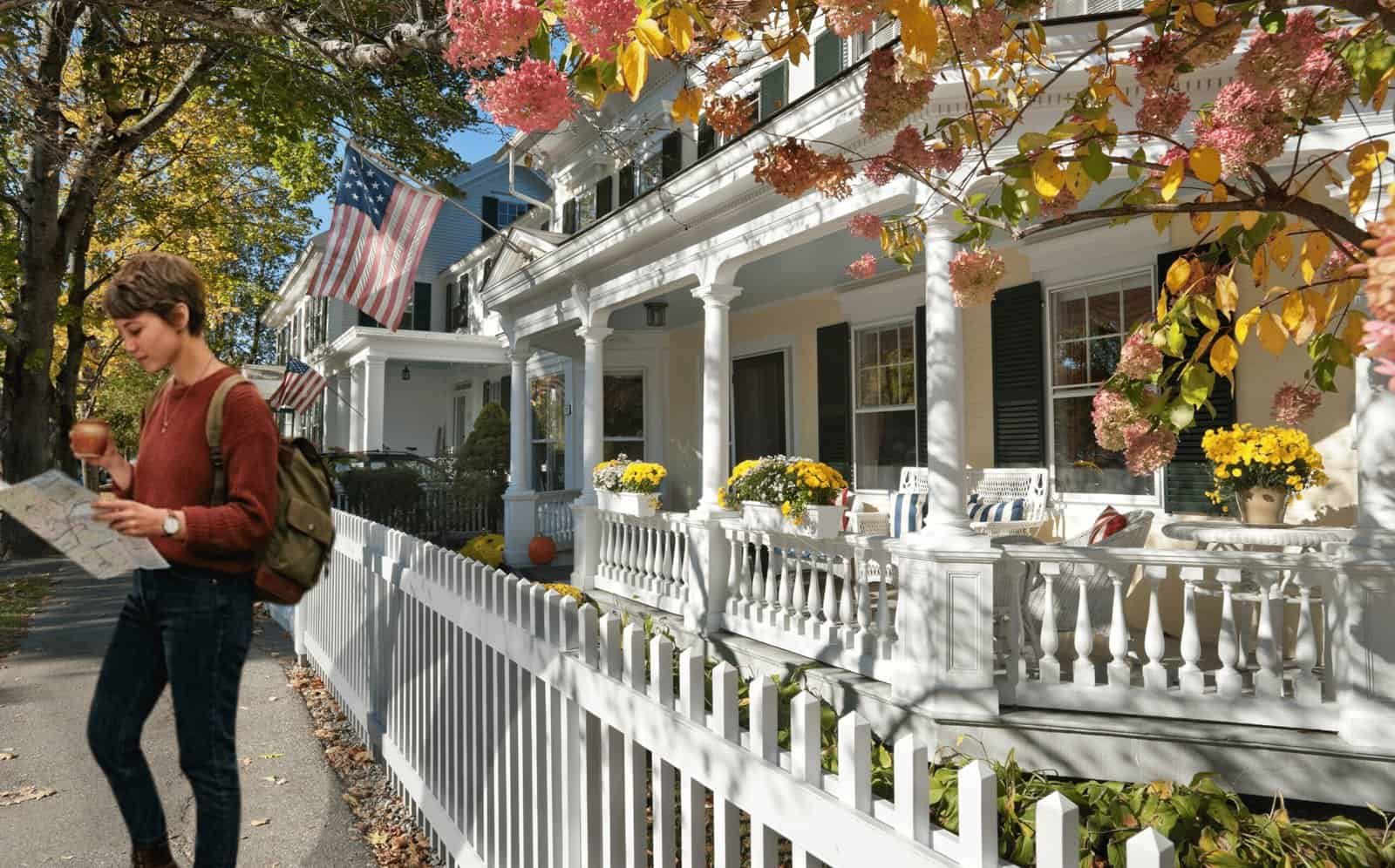 A young woman walking past a white historic house decorated with fall flowers and American flags, reading a map or brochure.