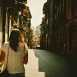 A woman walking down a quiet, sunlit city street with colorful buildings on either side, capturing the essence of slow travel and urban exploration.
