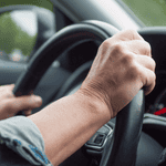 Close-up of a mature person's hand gripping a car steering wheel during a road trip, with a scenic view outside the window.