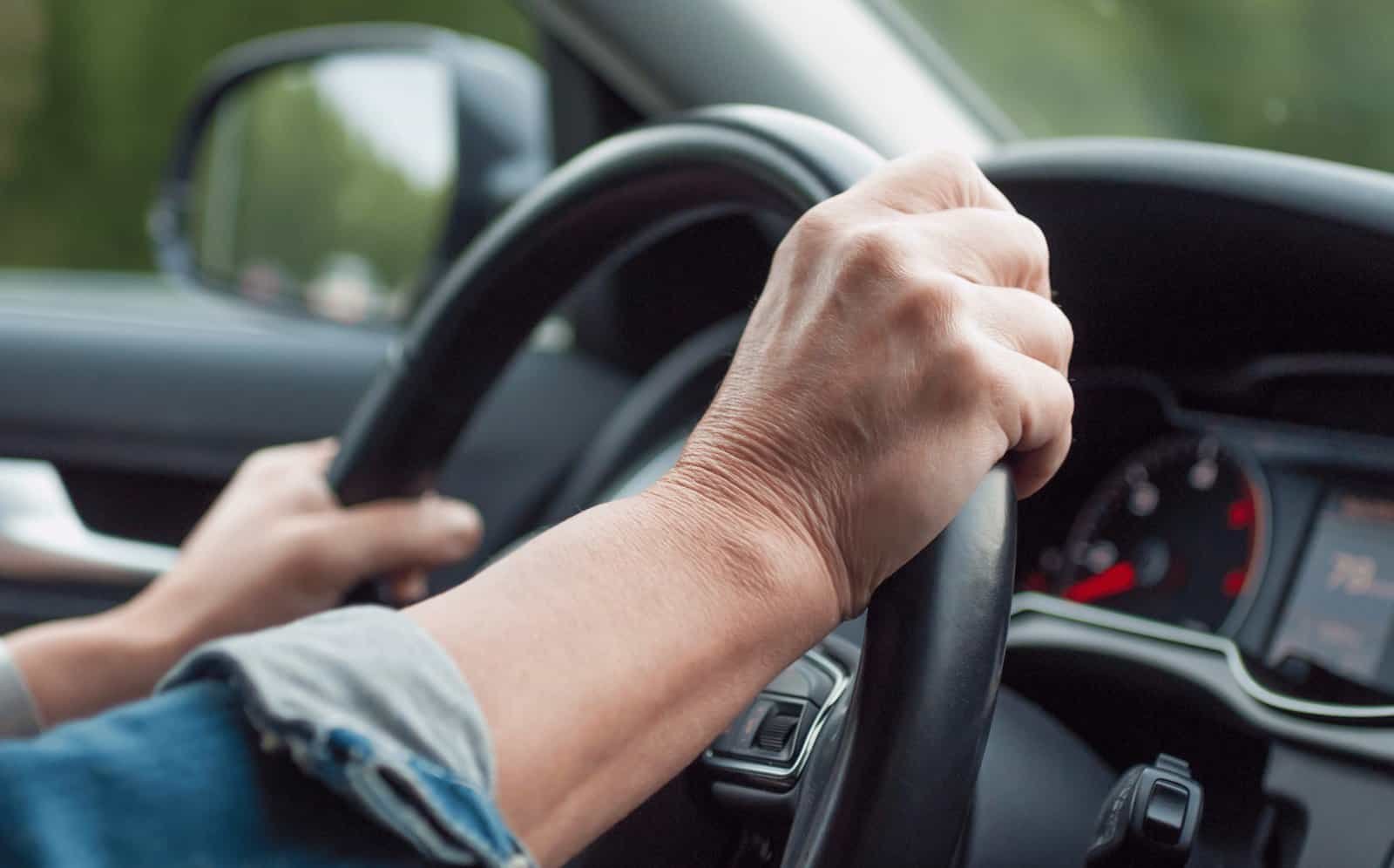 Close-up of a mature person's hand gripping a car steering wheel during a road trip, with a scenic view outside the window.